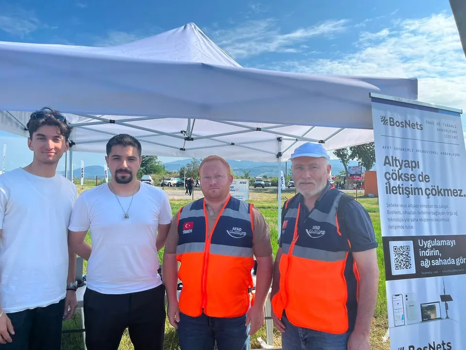 BosNets ekibinden iki kişi ve turuncu yelekli iki AFAD gönüllüsü stant önünde birlikte poz veriyor.